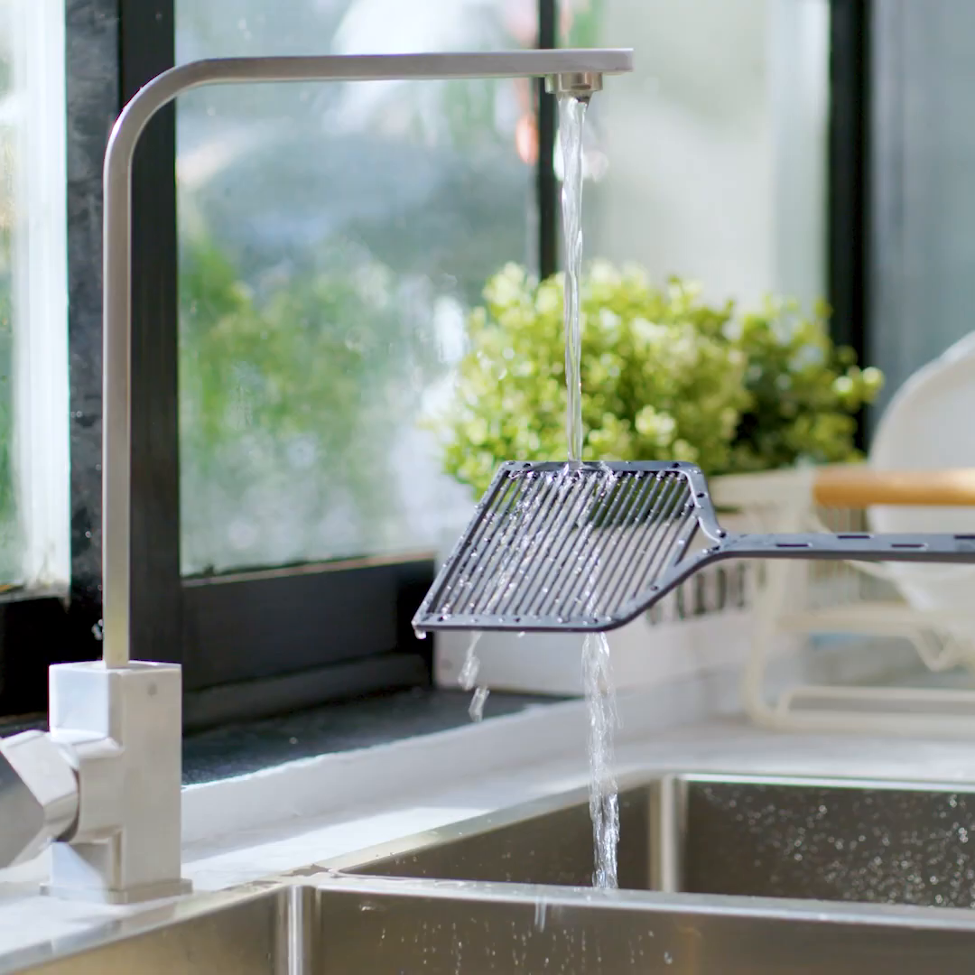 The square ZAPPER+ paddle being rinsed under a running kitchen tap, water flowing through the grid